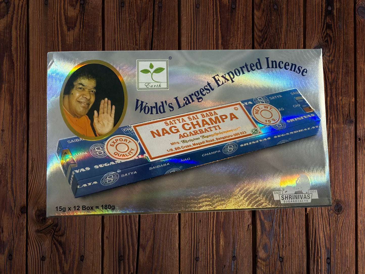 A 12-pack box of Srinivas Nag Champa Agarbatti incense sticks, displayed on a metallic holographic backing card against a dark wood background.