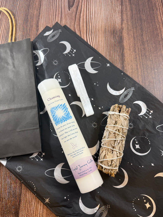 Cleansing pillar candle, selenite wand and white sage bundle on moon-patterned tissue beside a black gift bag