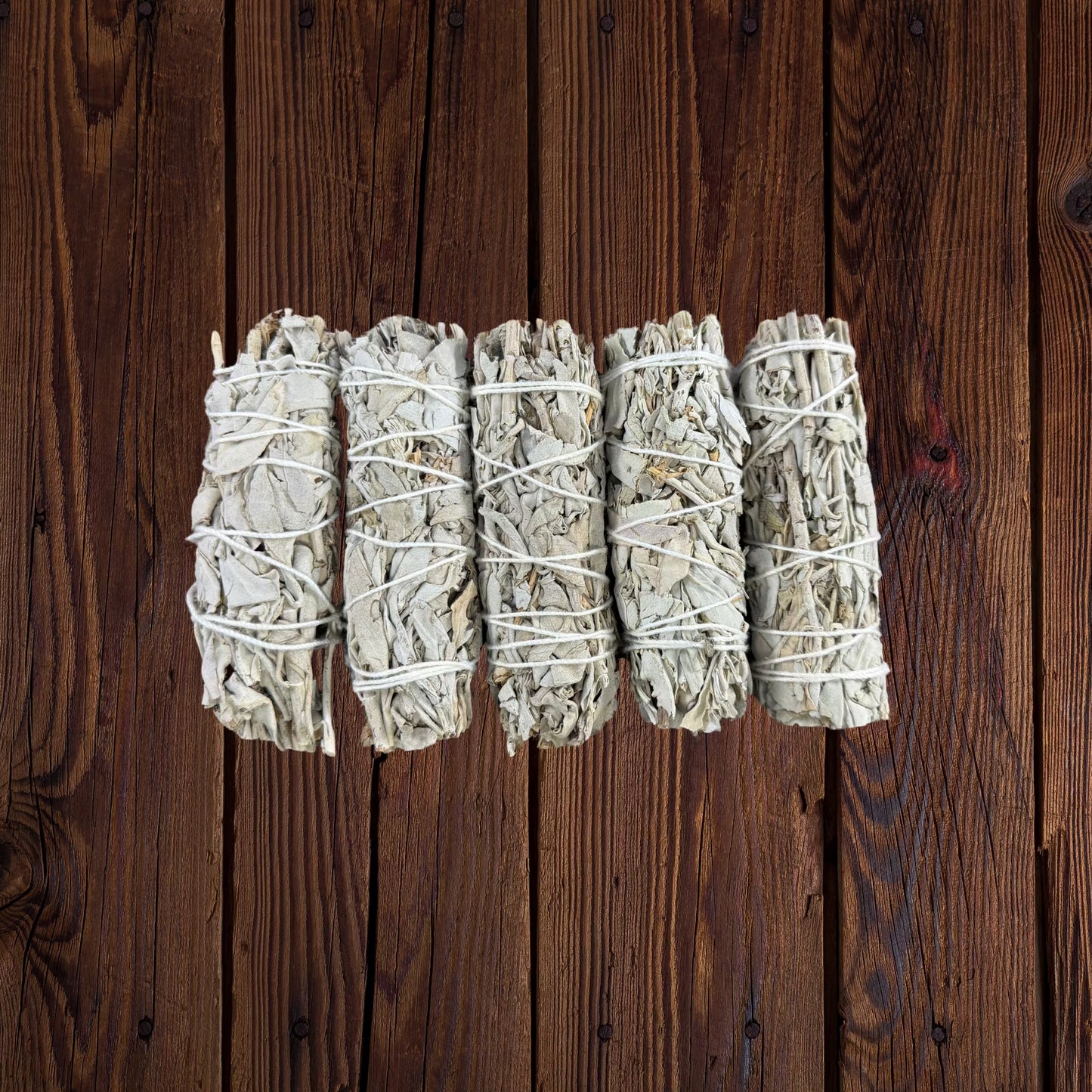 Five bundled white sage smudge sticks tied with string, arranged horizontally on a dark brown wooden surface.