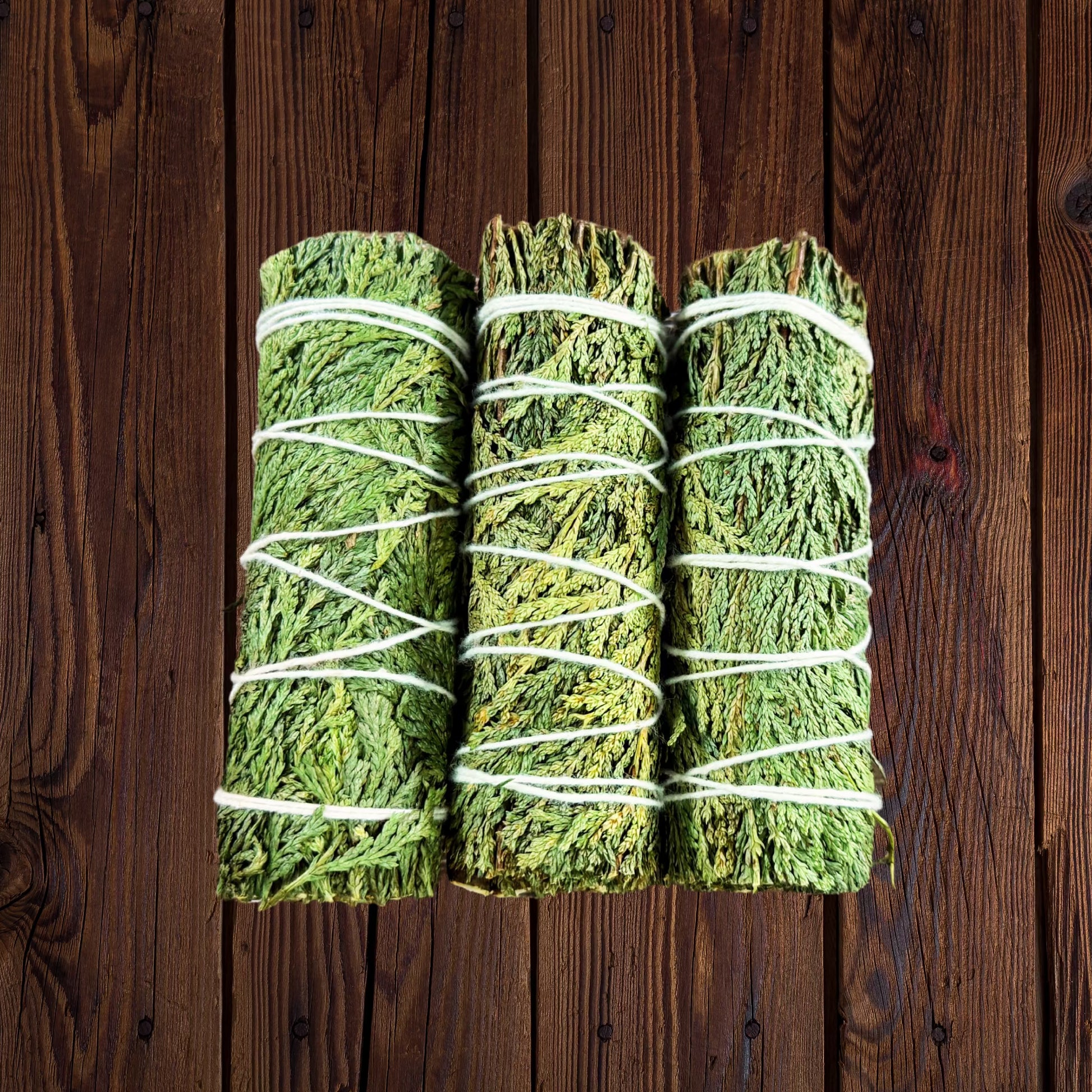 Three bundled green herbal or mossy sticks wrapped in white string, arranged vertically on a dark brown wood grain surface.