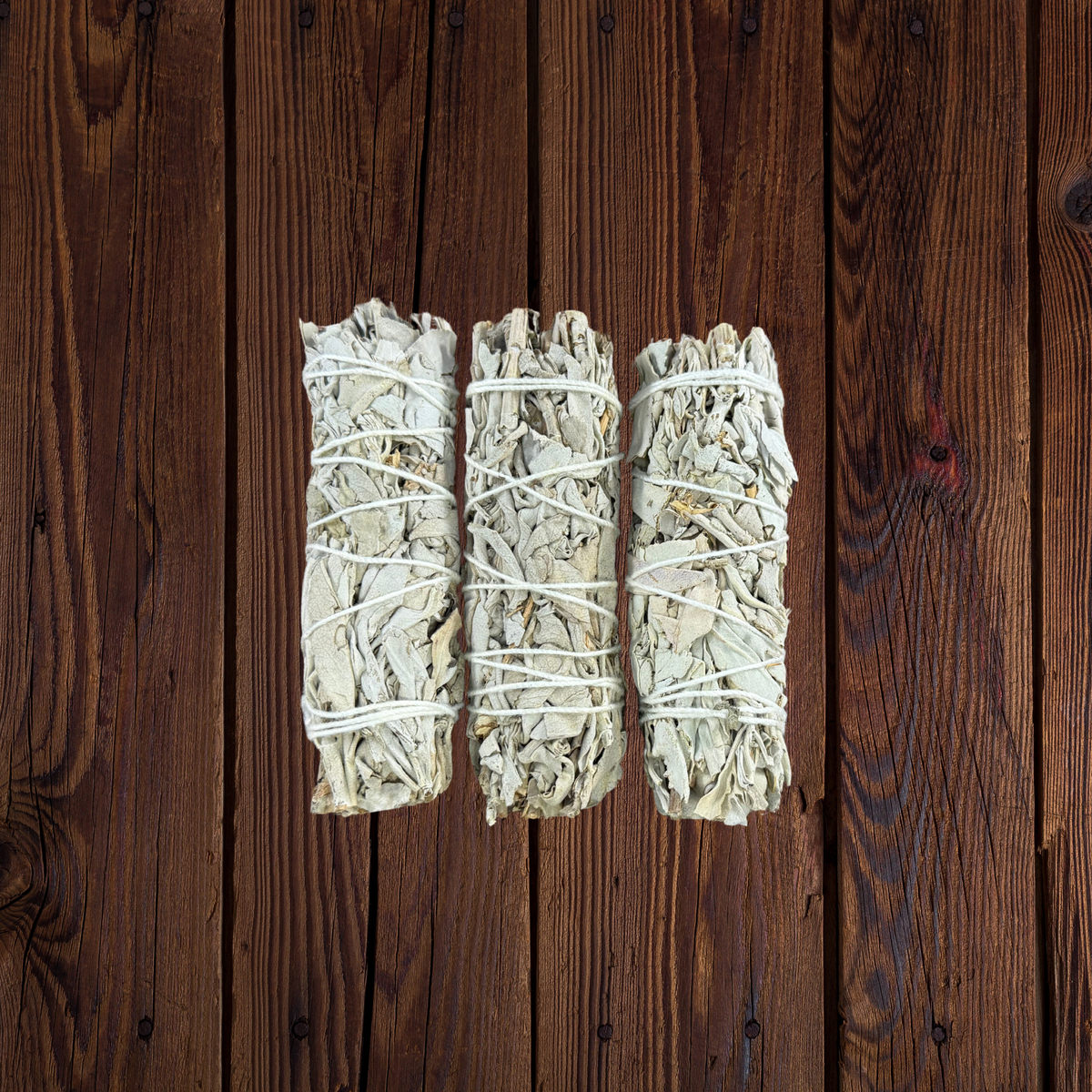 Three bundled white sage smudge sticks tied with white string, arranged vertically on a dark brown wooden plank background.