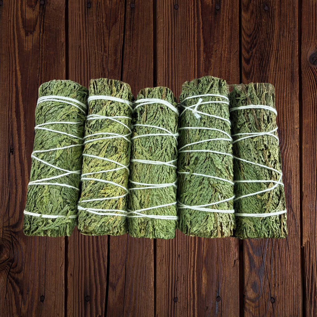 Five bundles of dried green herbs wrapped in white string, arranged side by side on a dark brown wooden plank background.